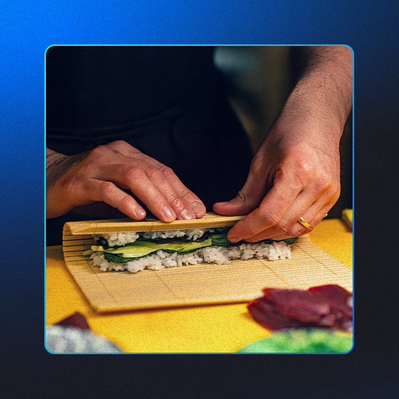 foto mostra um sushimaker preparando tipos de sushi diferentes, usando os ingredientes pepino, arroz e peixes.