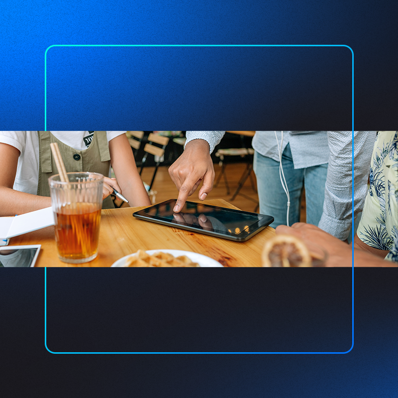foto mostra um zoom de mesa de restaurante com pratos, copos e 1 tablet com gestor de pedidos ifood aberto enquanto o atendente usa ele